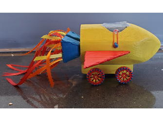 The side view of a spaceship-looking car. The body, painted yellow, is made out of wood. There's red cardboard wings on each side and blue cardboard boosters on the bottom. Red, orange, and yellow ribbons come out from it. There is a plastic cover, a red band wraps around a glob of hot glue painted blue. The wheels on the bottom are painted red, yellow, and blue.